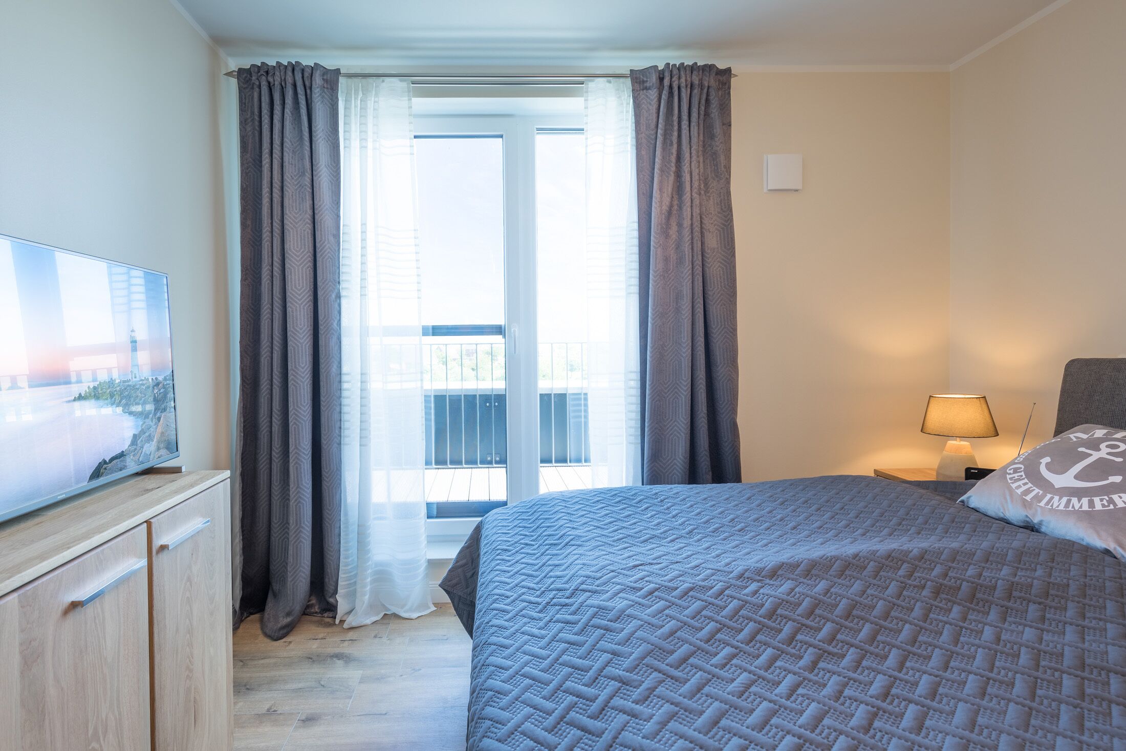 Ein Hotelzimmer mit Bett, Fernseher auf einer Holzkommode, Balkon mit Vorhängen und Lampe auf dem Nachttisch.