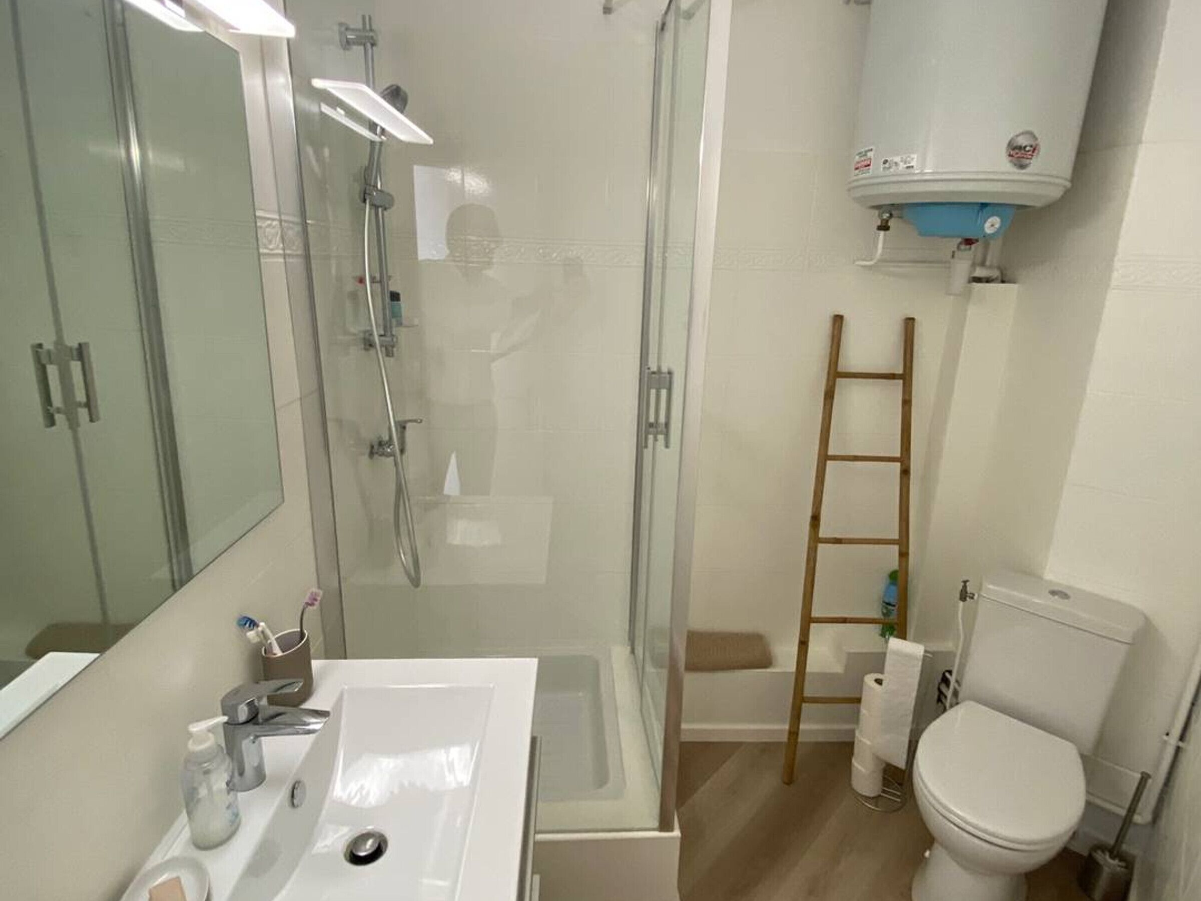 A bathroom with a glass shower enclosure, a white sink with a mirror, a toilet, and a ladder leaning against the wall.