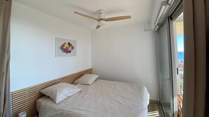 A bedroom with a bed, a ceiling fan, a window, and a view of the sea.