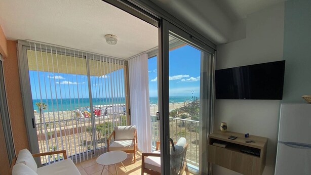 A balcony with a view of the beach, a sofa, a chair, a small table, and a TV mounted on the wall.