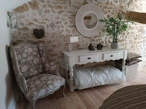 A room with a stone wall, a white console table with decorative items, a floral-patterned armchair, and a wooden floor. - "Magic Valley" Vacation Home in Panissaud (Le Grand-Madieu)
