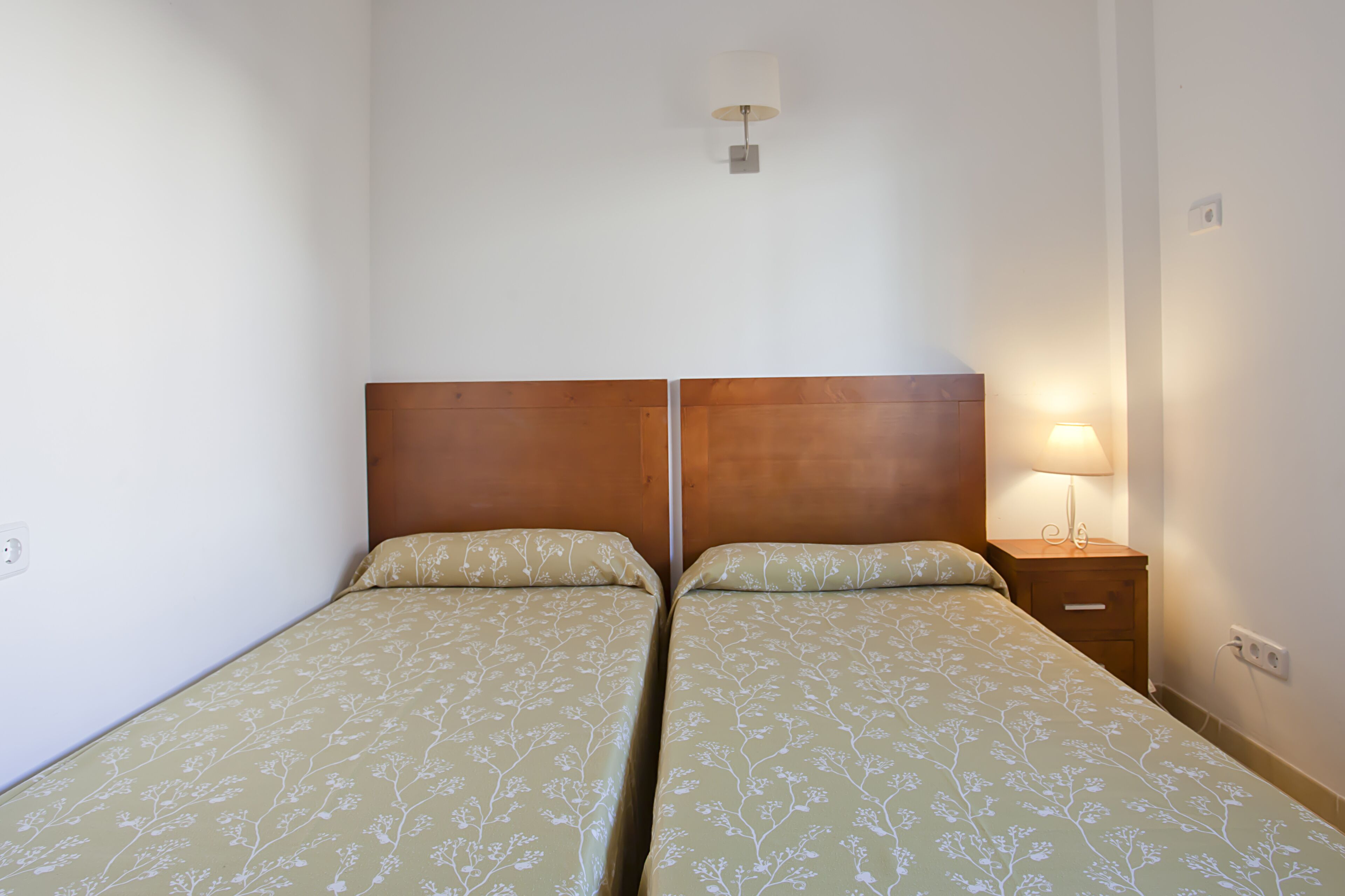 A room with two beds, a nightstand, and a wall-mounted lamp.