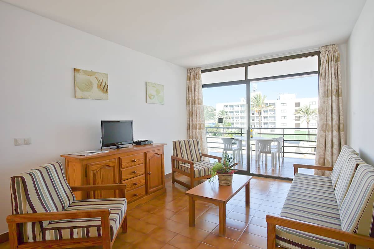 Una sala de estar con un sofá, sillones, un televisor y un mueble de madera. Hay un balcón con vistas a edificios y palmeras.
