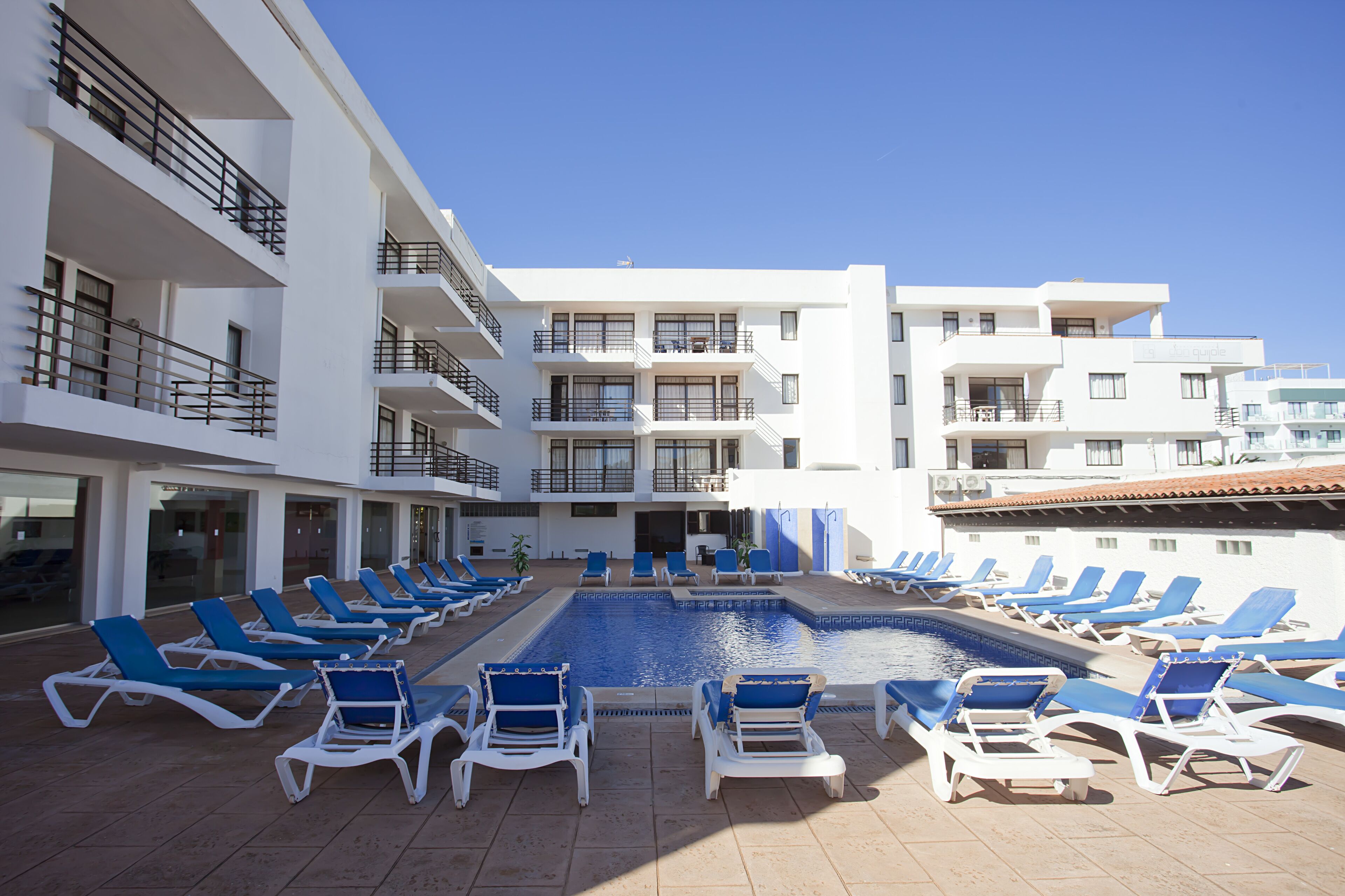 A poolside area with sun loungers and a swimming pool, surrounded by multi-story buildings with balconies.