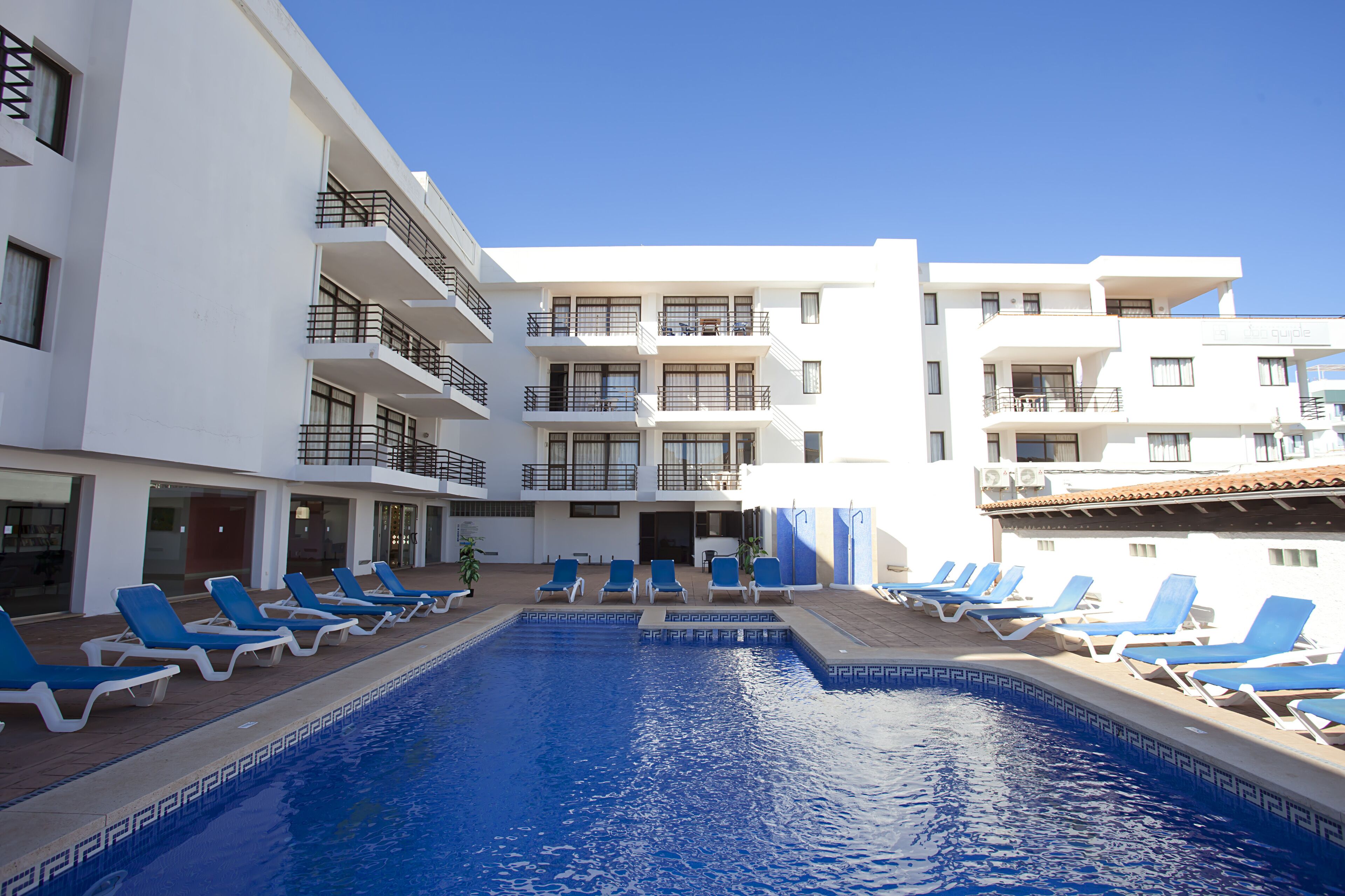 A swimming pool with blue lounge chairs and a white building with balconies.