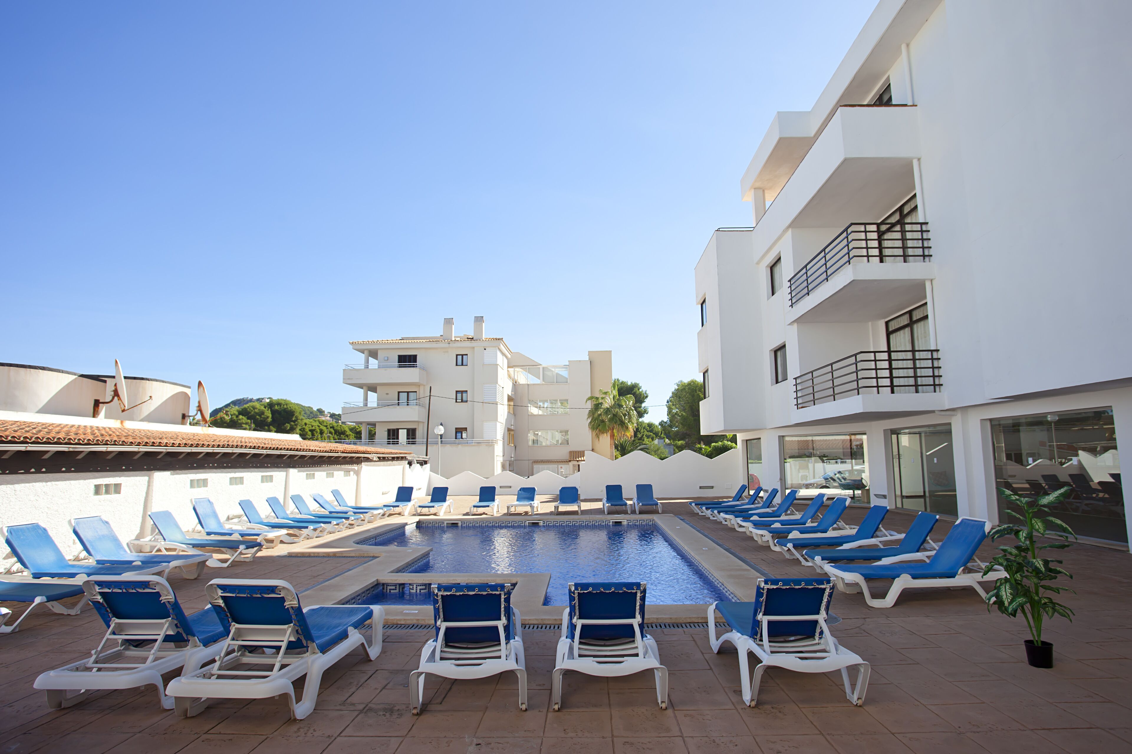 A poolside area with lounge chairs arranged around a central pool, adjacent to a white building with balconies.