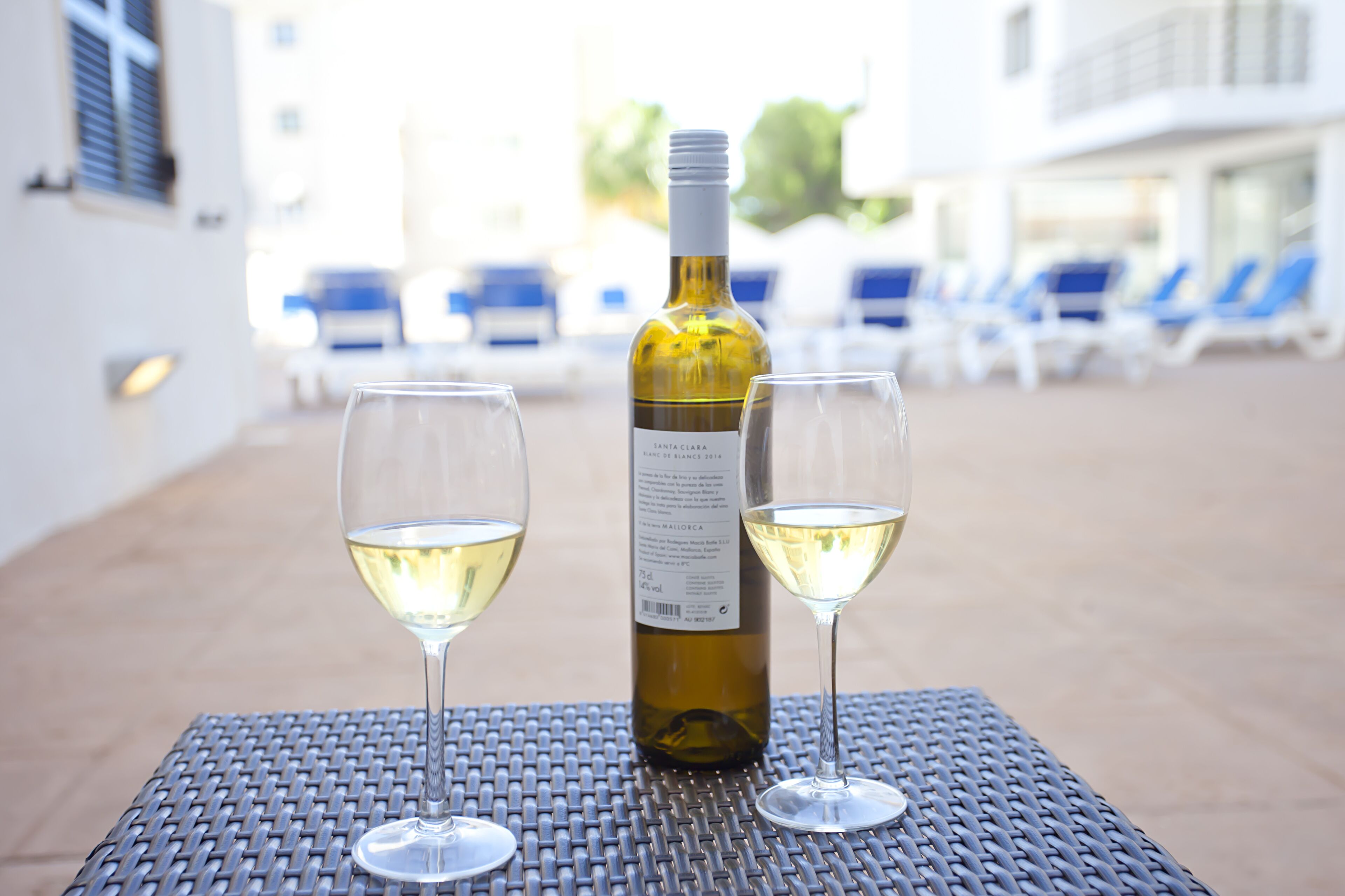 A bottle of wine and two glasses on a table in an outdoor setting with lounge chairs in the background.