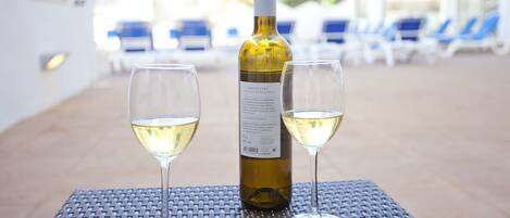 A bottle of wine and two glasses on a table in an outdoor setting with lounge chairs in the background.