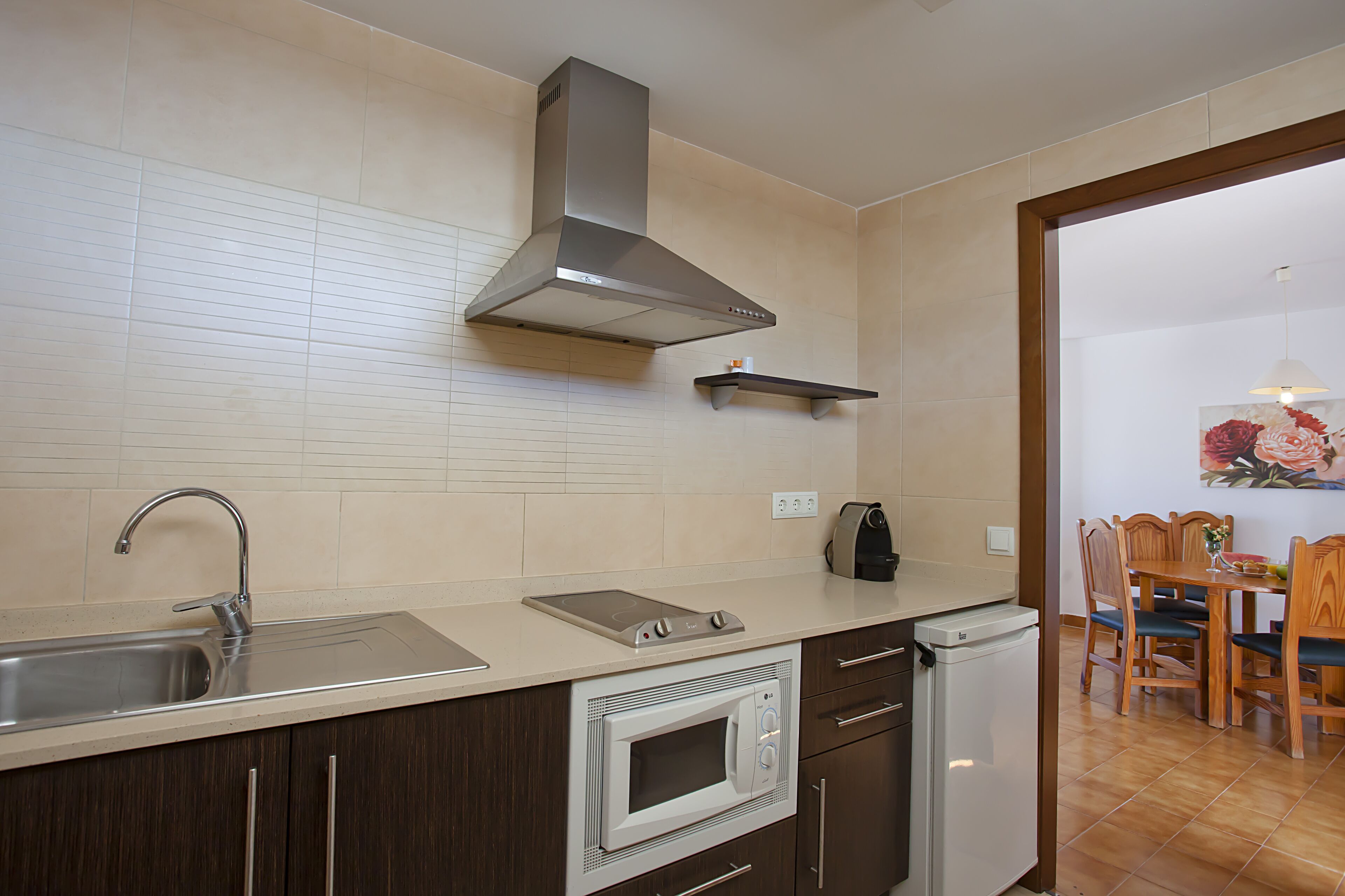 A kitchen with a stainless steel sink, a microwave, and a built-in oven. There is a wooden dining table with chairs and a hanging light fixture.