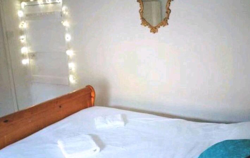 Ein Bett mit weißen Leinen, ein hölzernes Kopfbrett, ein Spiegel mit goldener Rahmung und eine Lichterkette an der Wand.