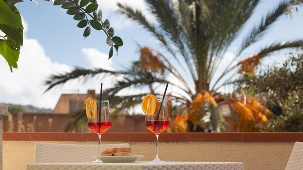 Deux verres d’une boisson rouge, garnis de tranches d’orange, sont posés sur une table devant une chaise blanche, le tout se détachant sur un fond de palmiers et d’un ciel dégagé.