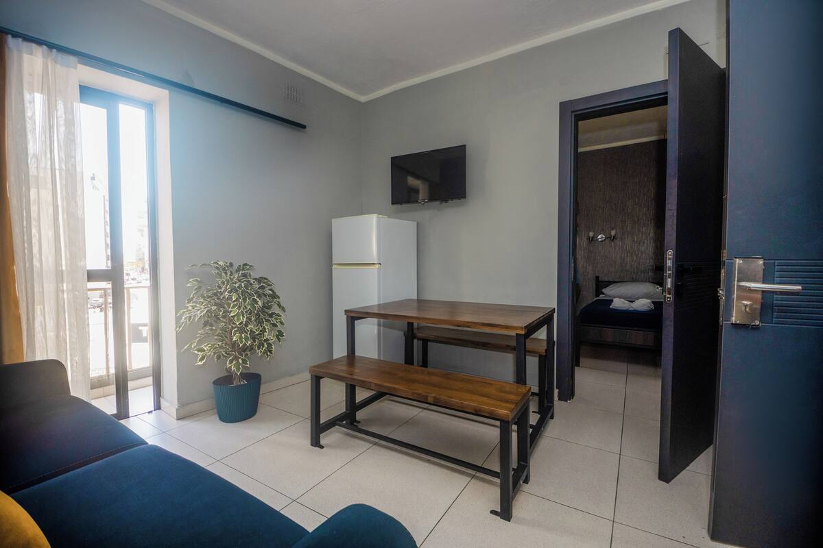 Un espace de vie compact comprenant un canapé, une table à manger, un banc, une plante en pot, un réfrigérateur, un téléviseur et une porte donnant sur une chambre.