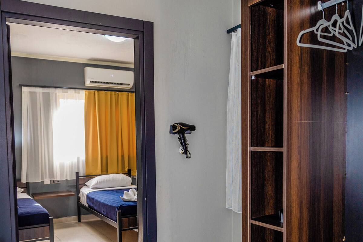 Une chambre d’hôtel de taille modeste, comprenant un lit simple, une armoire en bois, un petit bureau et une plante en pot.