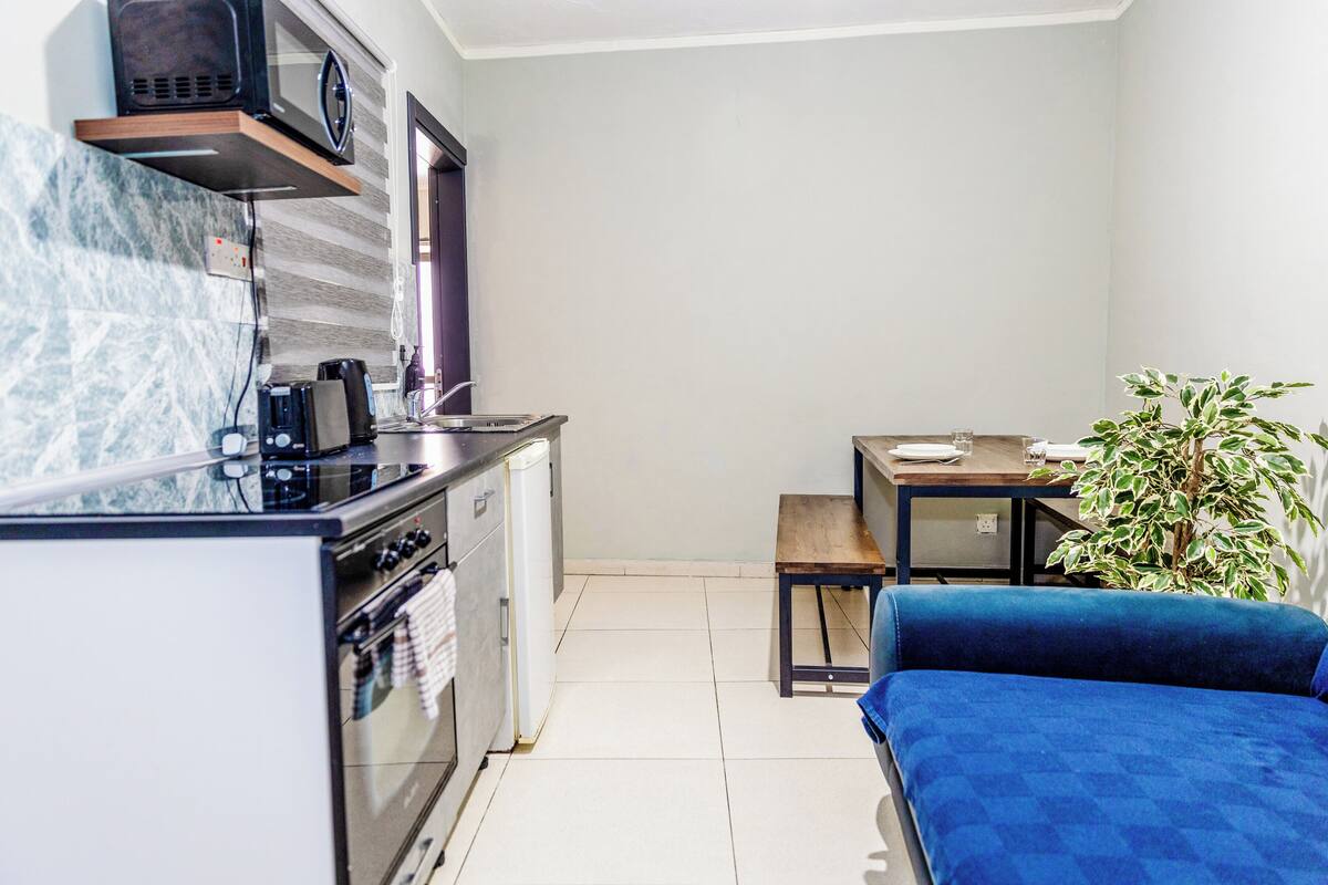 Une cuisine compacte équipée d’un four à micro-ondes, d’un four et d’une machine à café. On y trouve une table à manger avec des chaises et un petit canapé bleu. Une plante en pot est visible dans un coin.
