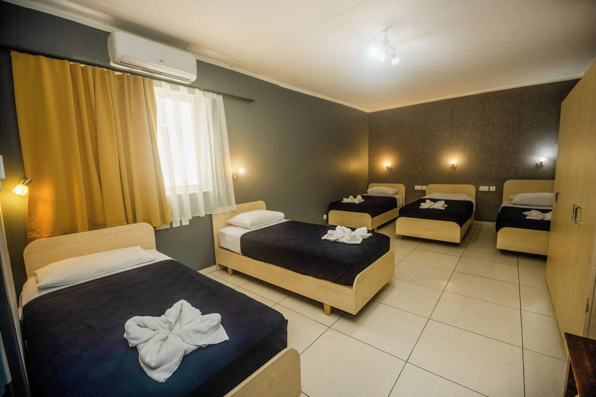 Une chambre d’hôtel avec quatre lits, une grande fenêtre avec des rideaux, un appareil de climatisation et des lampes fixées au mur.