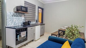 A compact kitchen with a microwave, oven, and sink, a blue sofa with a yellow pillow, and a potted plant in a small dining area. - The Seven Apartments (St. Julian's)