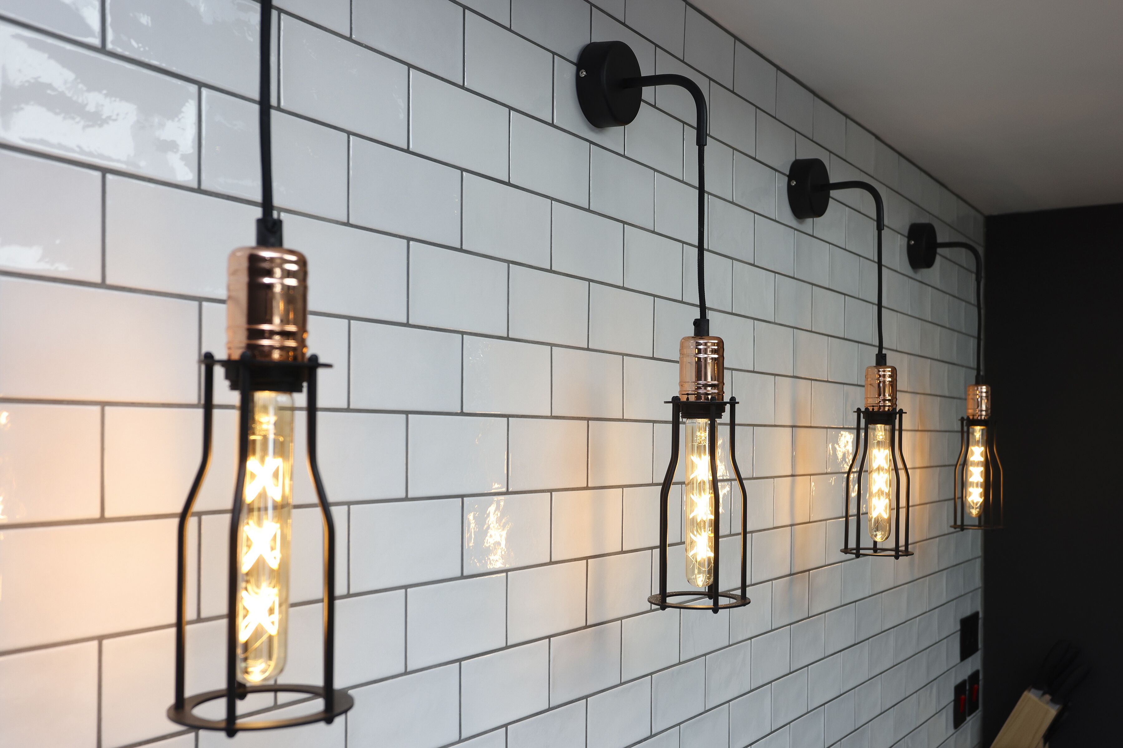 A wall with white tiles and four hanging light fixtures with exposed bulbs.