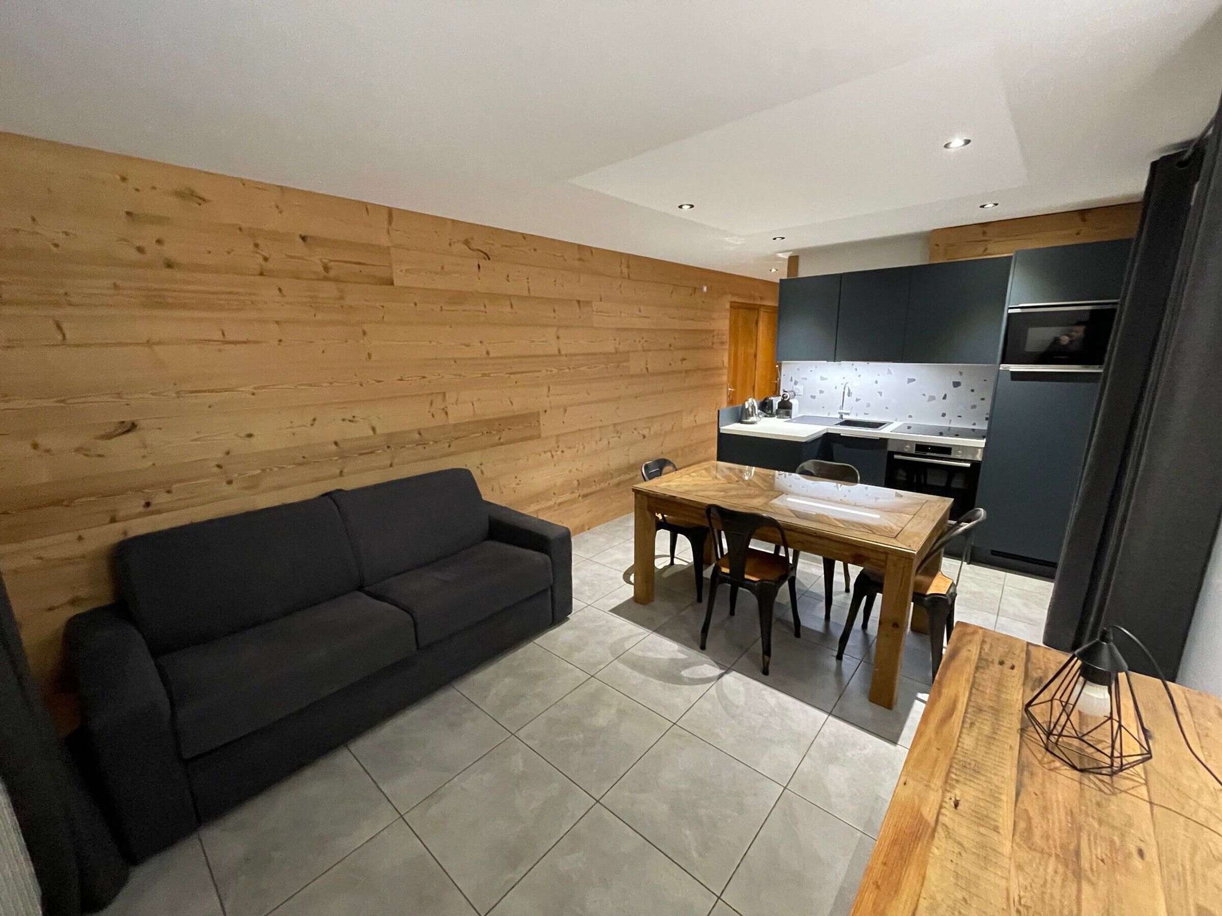 Una sala de estar moderna con pared de madera, un sofá negro, una mesa de comedor con sillas y una cocina con armarios oscuros y un microondas.