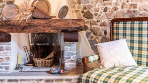 Une pièce chaleureuse avec une cheminée en pierre, un lit à carreaux et une étagère en bois où sont disposés divers objets.