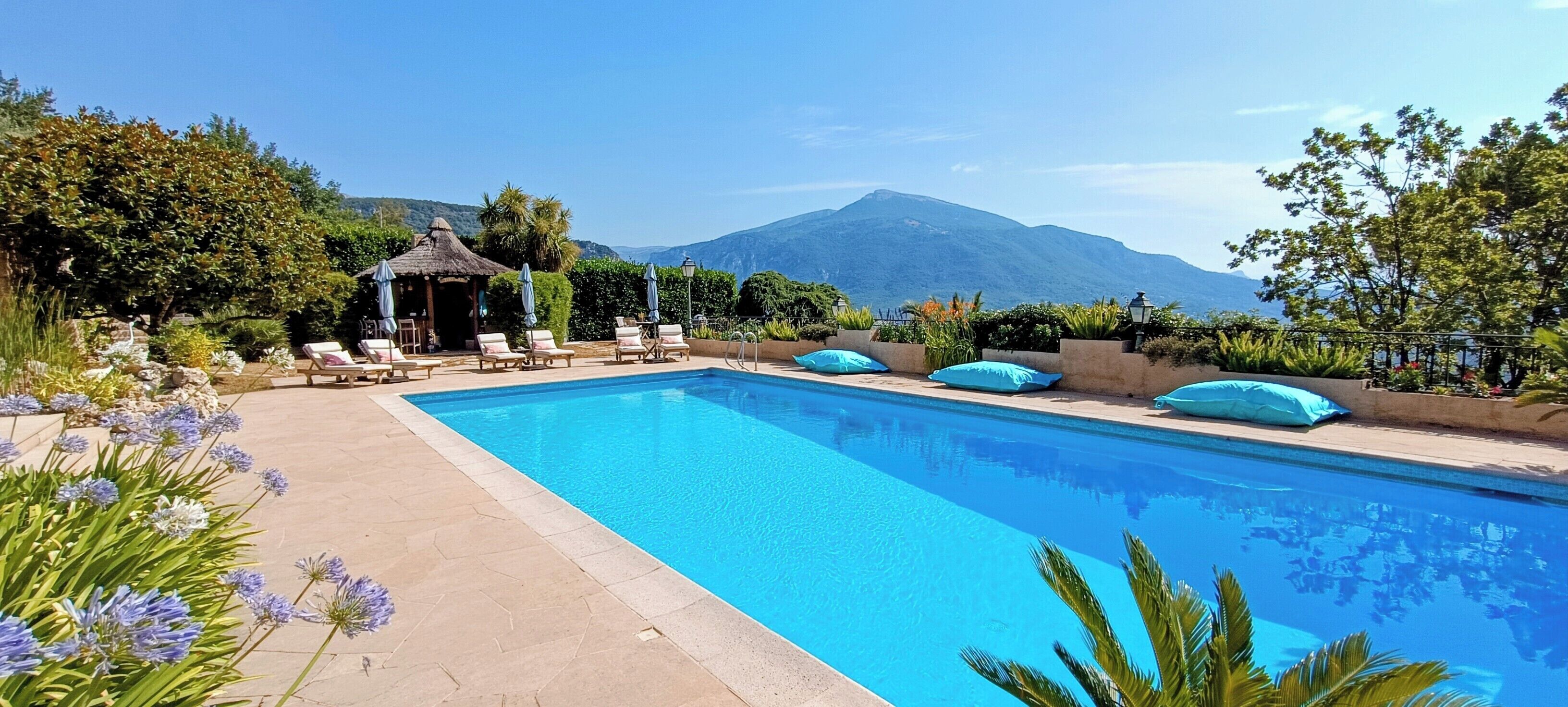 A rectangular pool with lounge chairs and umbrellas, surrounded by greenery and a mountain view.