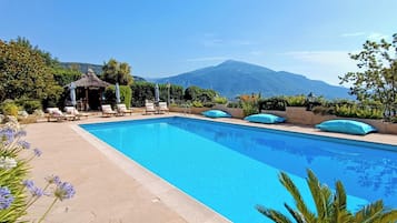 A rectangular pool with lounge chairs and umbrellas, surrounded by greenery and a mountain view.