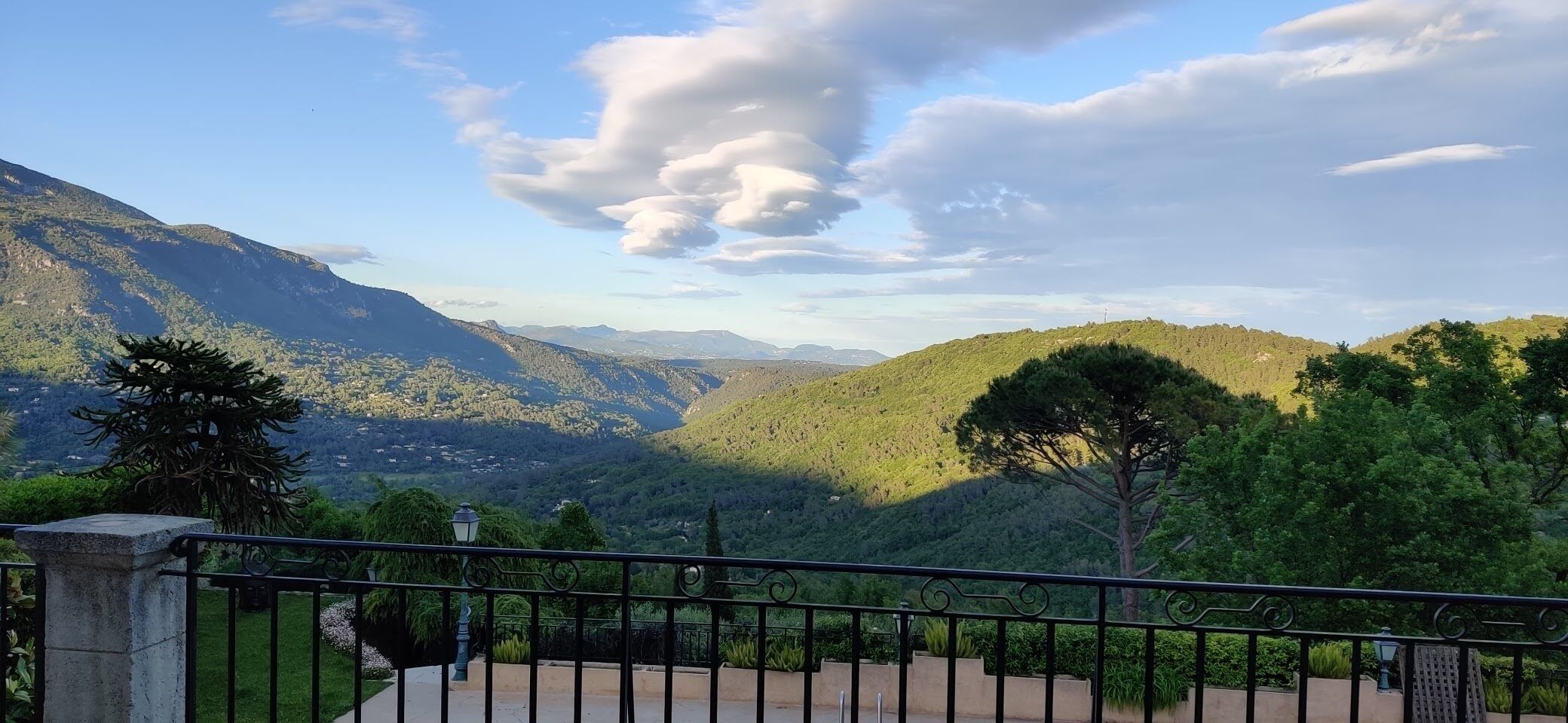Une vue panoramique depuis un balcon doté d’une balustrade, donnant sur une vallée bordée de montagnes et d’arbres.