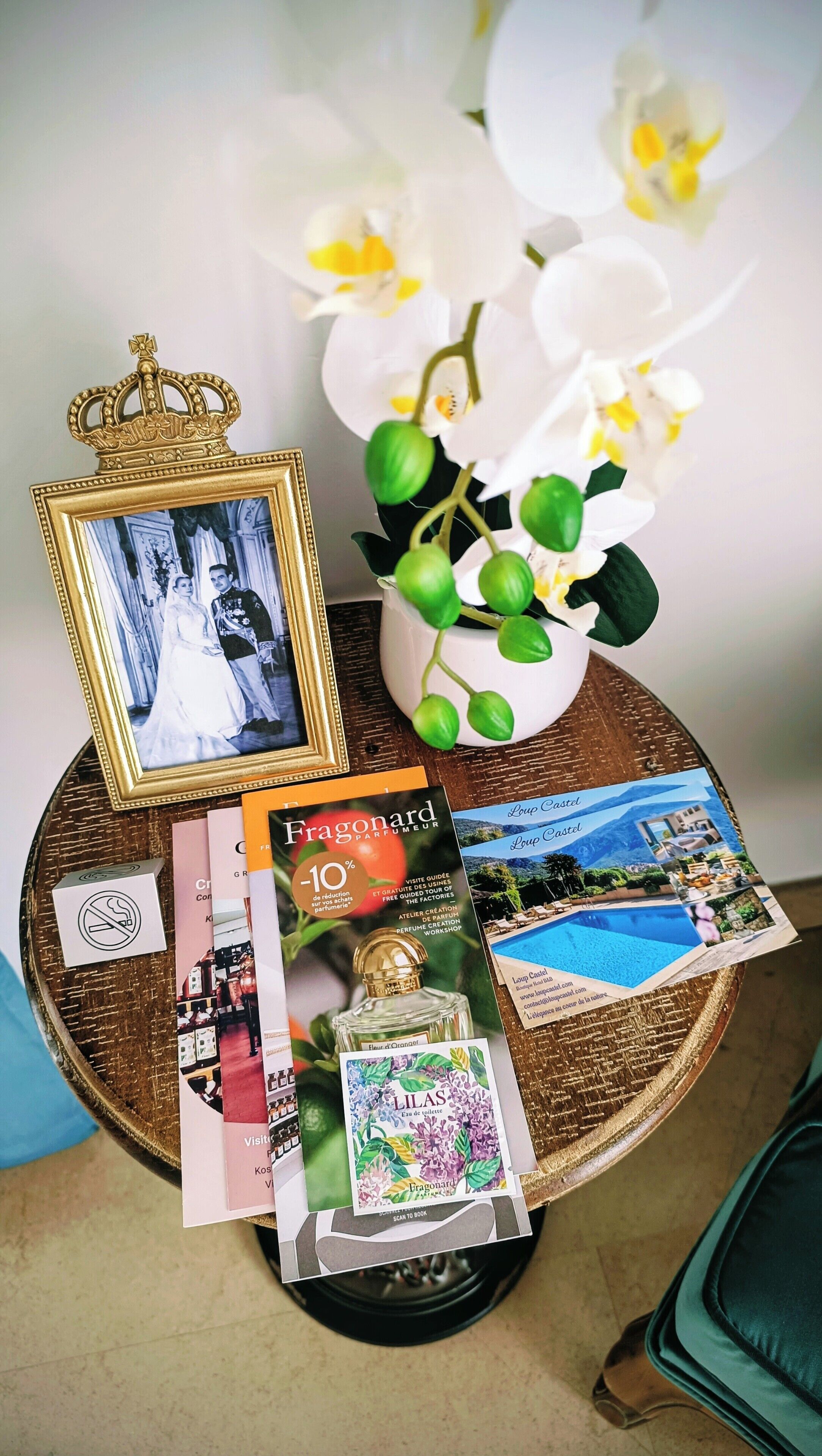 A round table with a framed photograph, a perfume bottle, brochures, and a vase with white flowers.