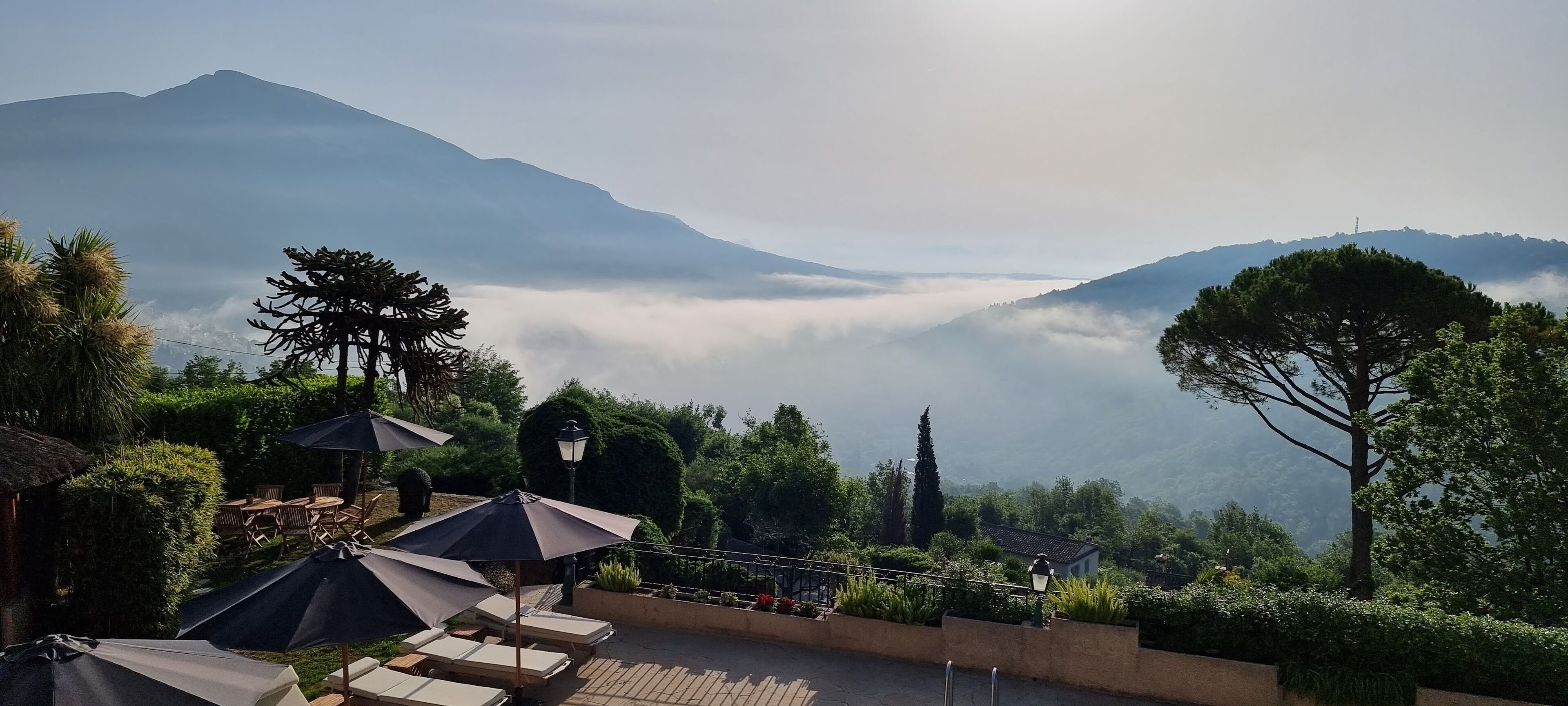 A terrace with lounge chairs and umbrellas overlooking a mountainous landscape with a valley shrouded in mist.