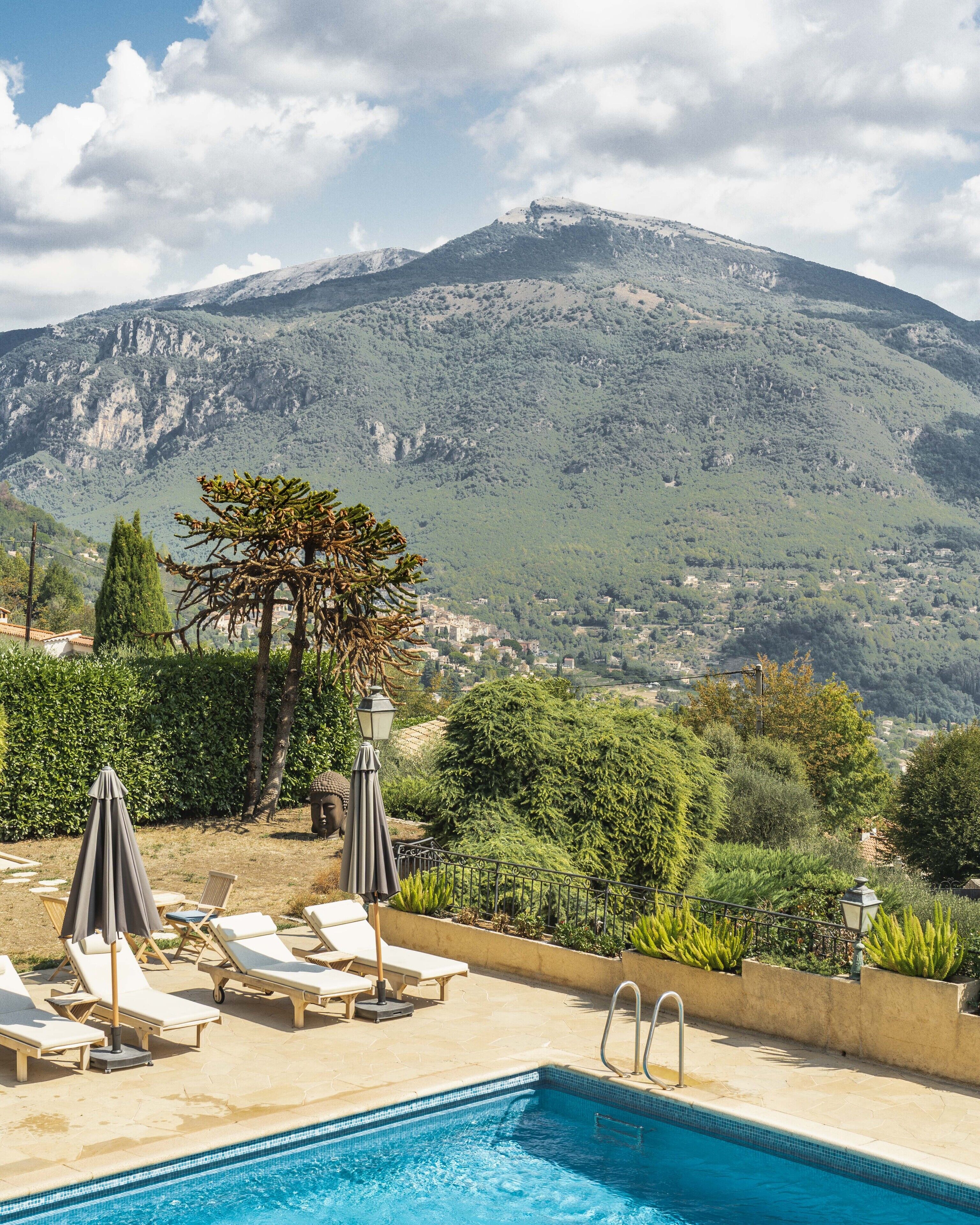 Una piscina con sillas de descanso y sombrillas, rodeada de vegetación y montañas al fondo.