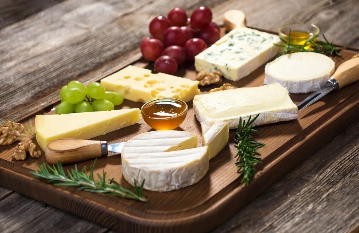 A wooden board with various cheeses, grapes, walnuts, and a small bowl of honey.