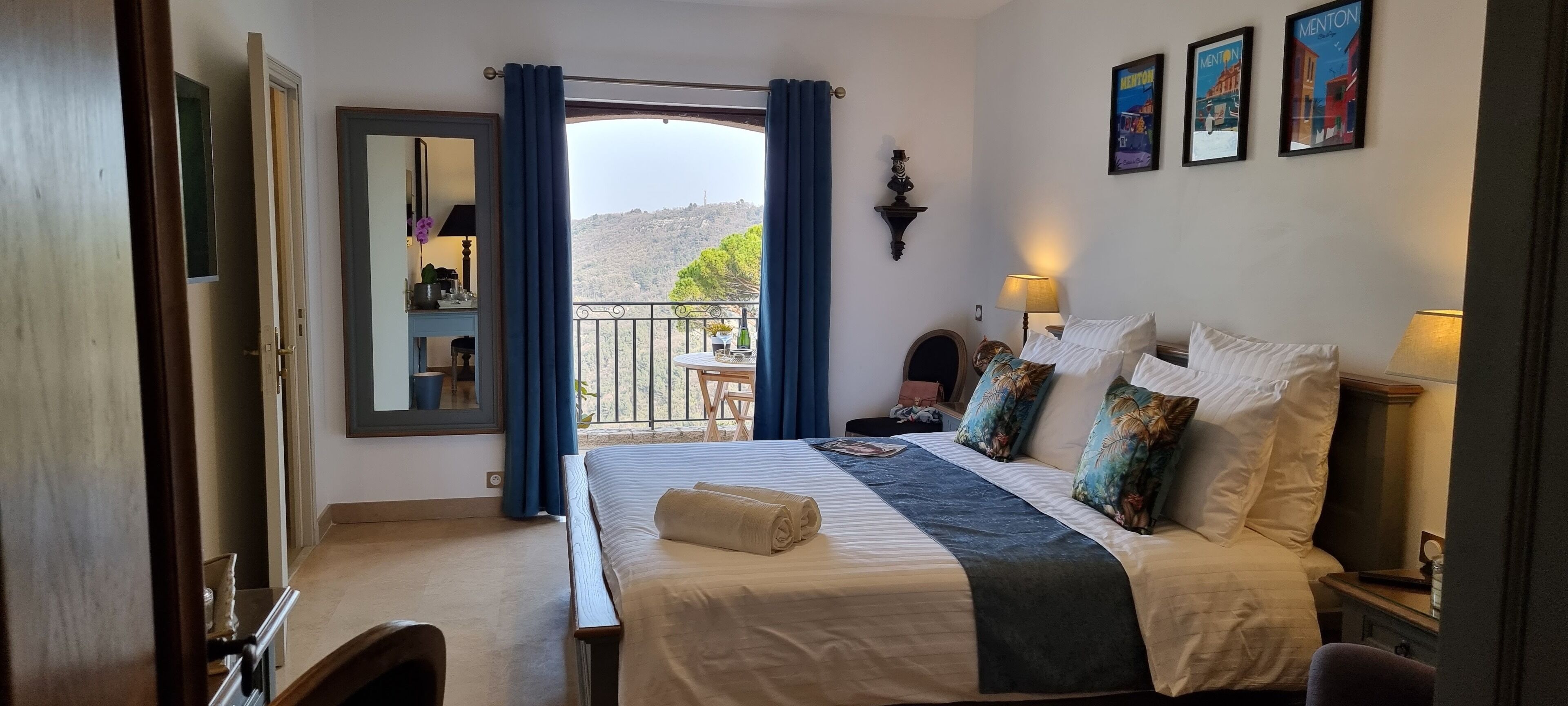 Ein Hotelzimmer mit Bett, Nachttischen, einem Balkon mit Tisch und Stühlen sowie Blick auf eine hügelige Landschaft.