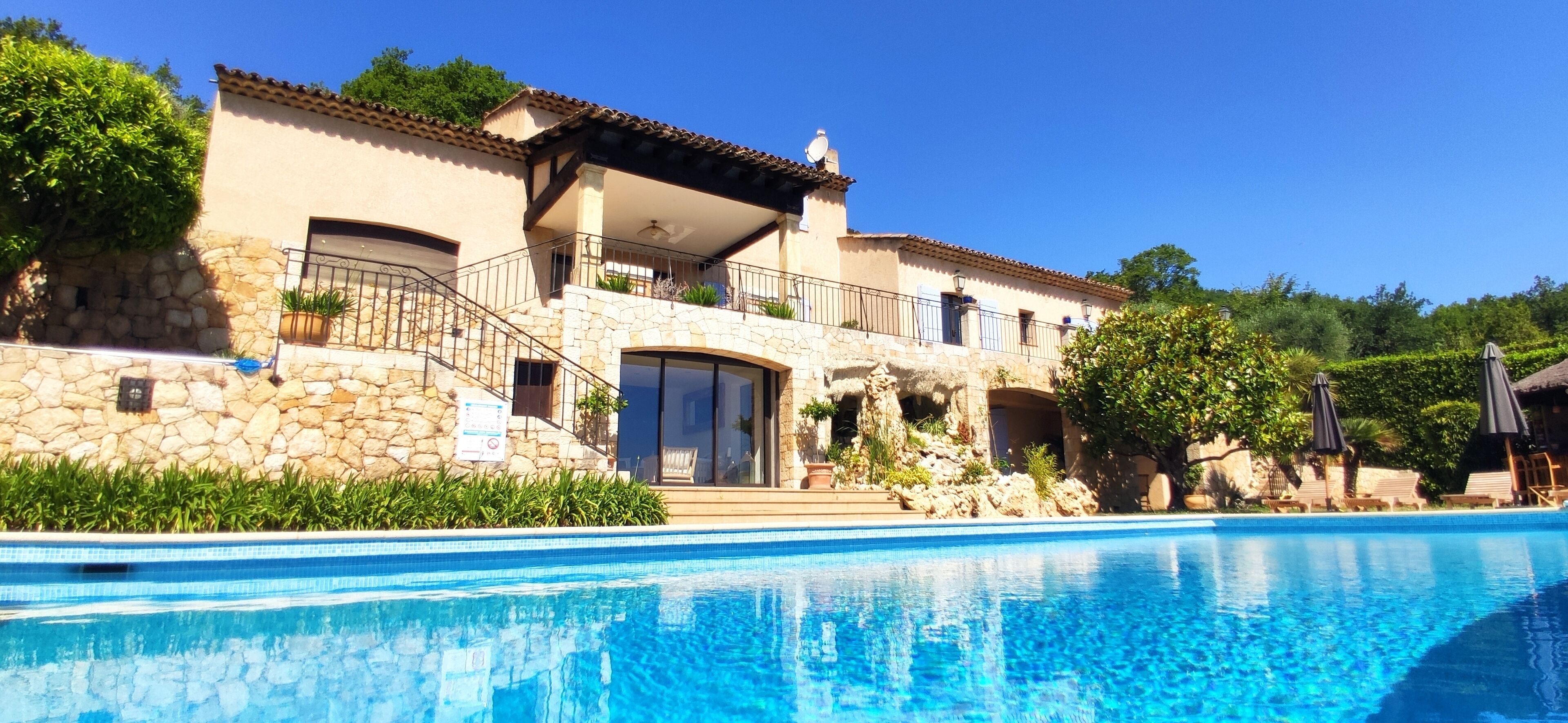 Vista a la piscina de una villa de estilo mediterráneo con fachada de piedra, un balcón con mobiliario exterior y una piscina azul cristalina.