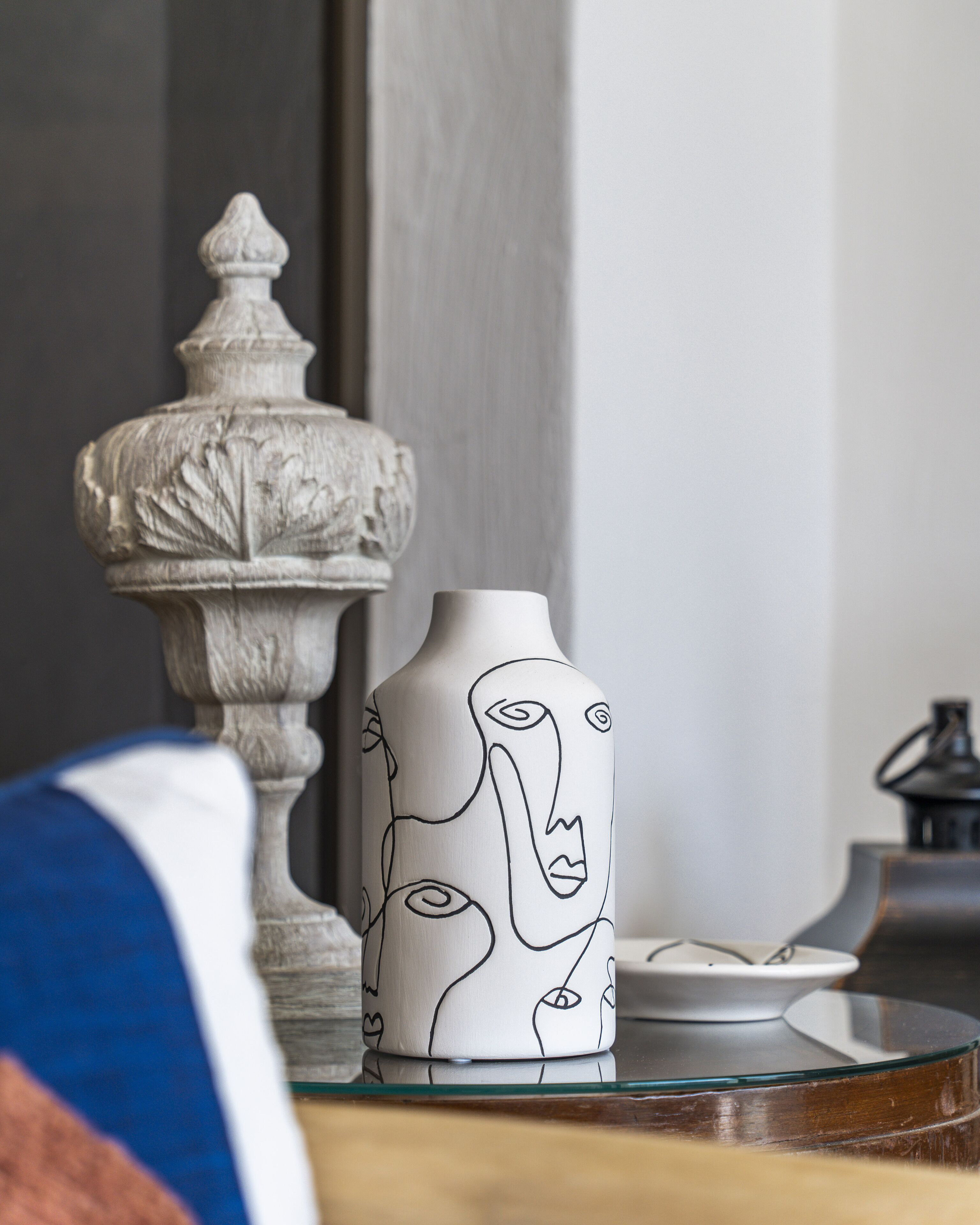 A white vase with abstract line art on a glass table, next to a decorative stone urn.