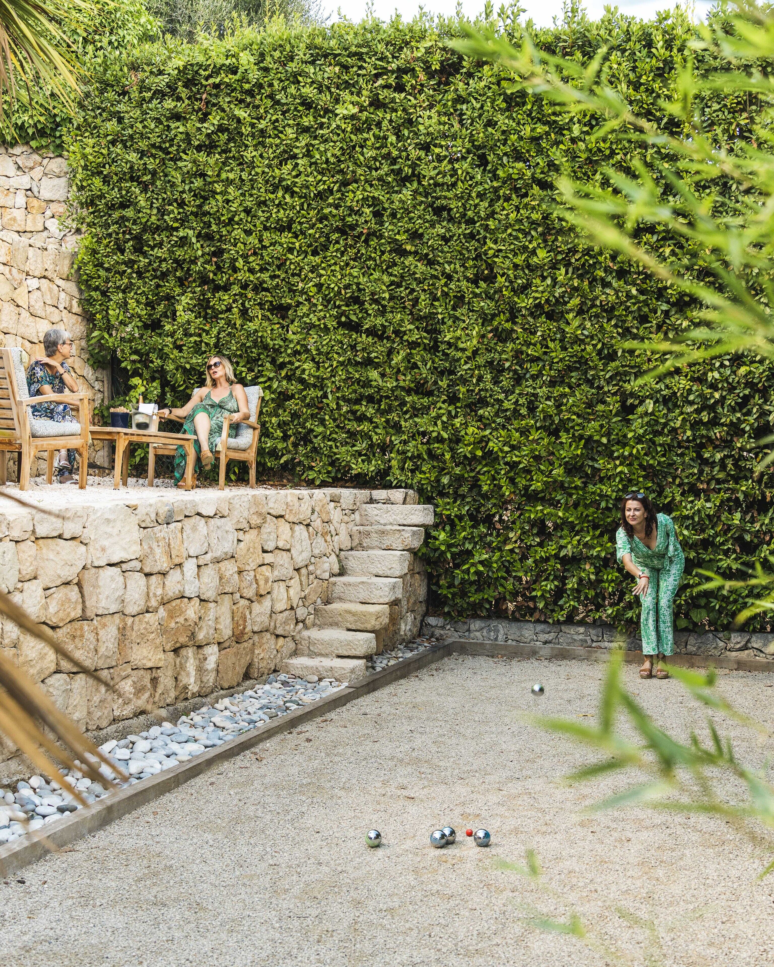 A stone wall with a hedge behind it, a gravel area with a few scattered balls, and two people sitting on chairs.