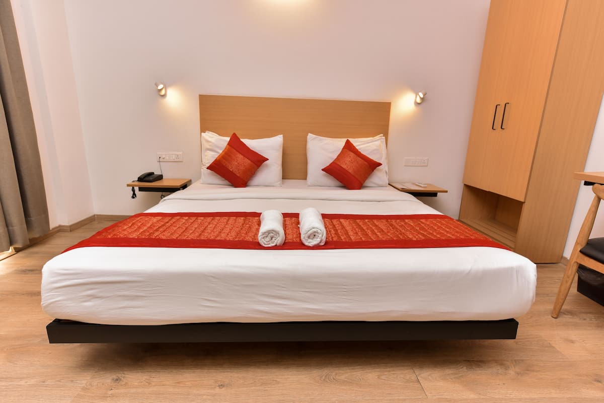A neatly made bed with a red and white bedspread, two red pillows, and two rolled towels. There is a wooden headboard, bedside tables with lamps, and a wooden wardrobe.