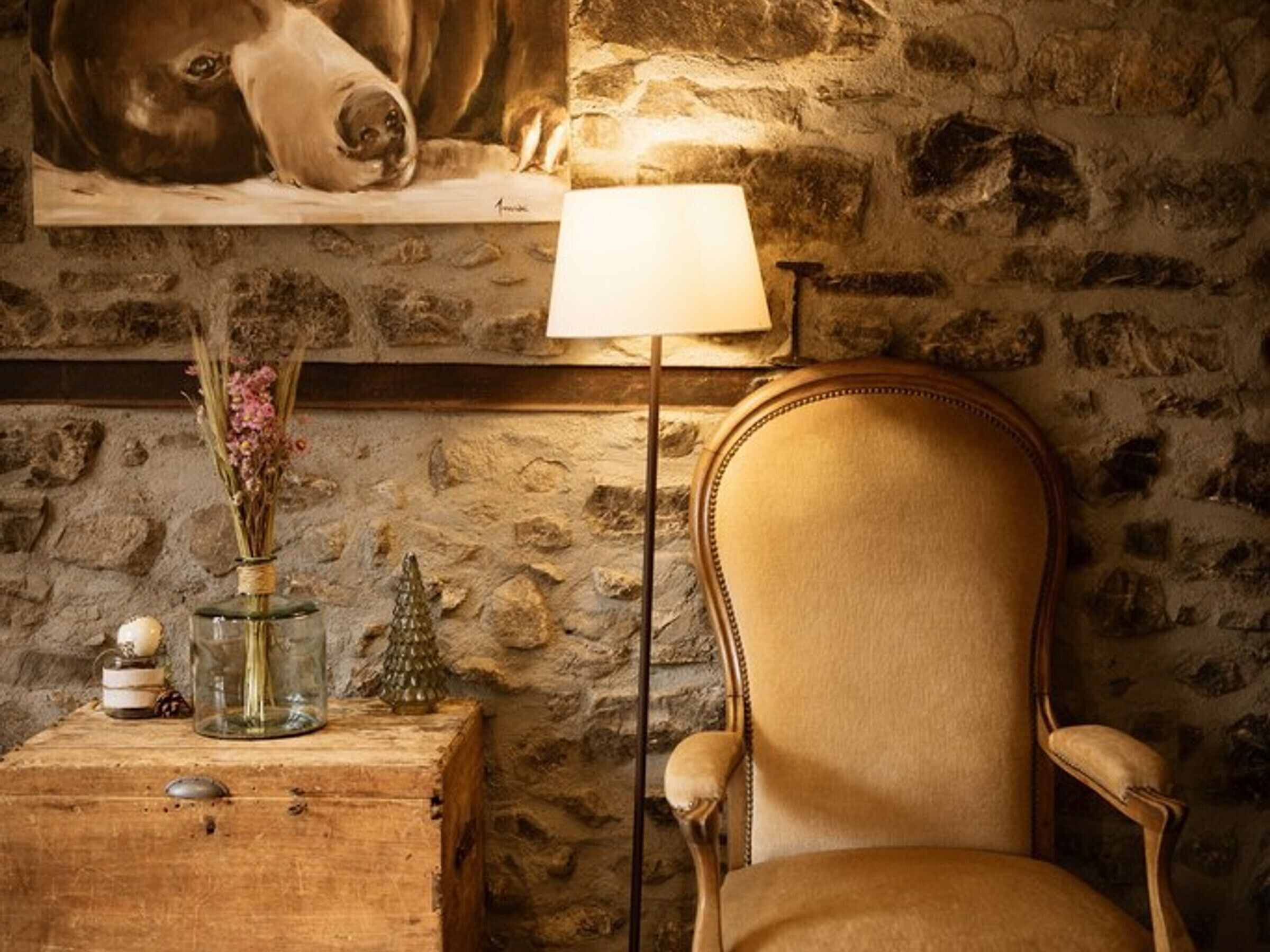 A rustic room with a stone wall, a wooden table with a lamp, a chair, and a vase with flowers.