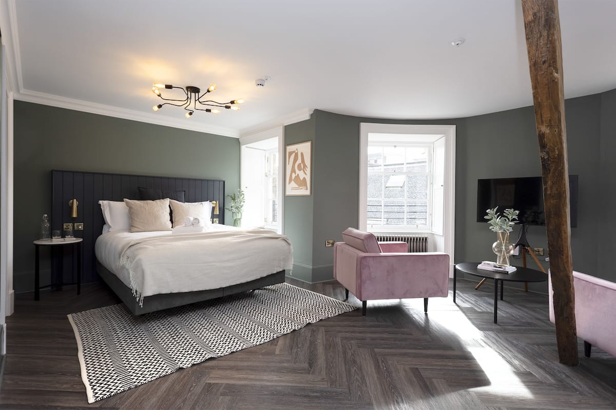 A modern bedroom with a large bed, a pink chair, a small table with a vase, and a large window.