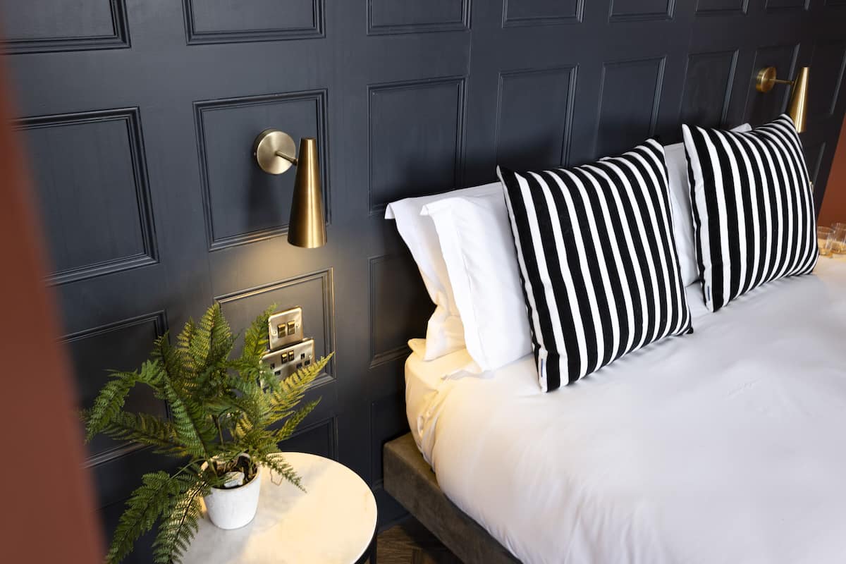 A neatly made bed with striped pillows, a bedside plant, and a wall-mounted light fixture.
