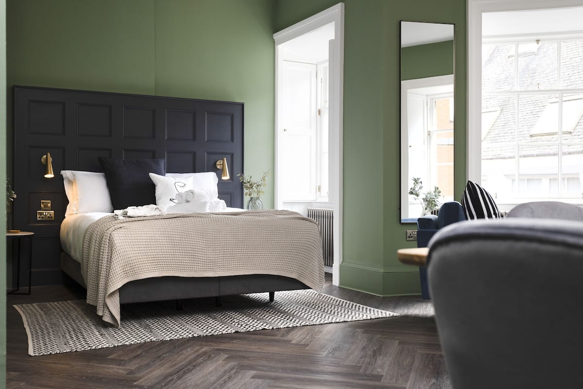 A bedroom with a dark headboard, a bed with white and beige bedding, a patterned rug, and a view of the outside through a window.