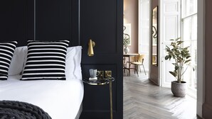 Une chambre avec un lit, des oreillers à rayures noires et blanches, une petite table de chevet avec une lampe, et une plante en pot près de la fenêtre.