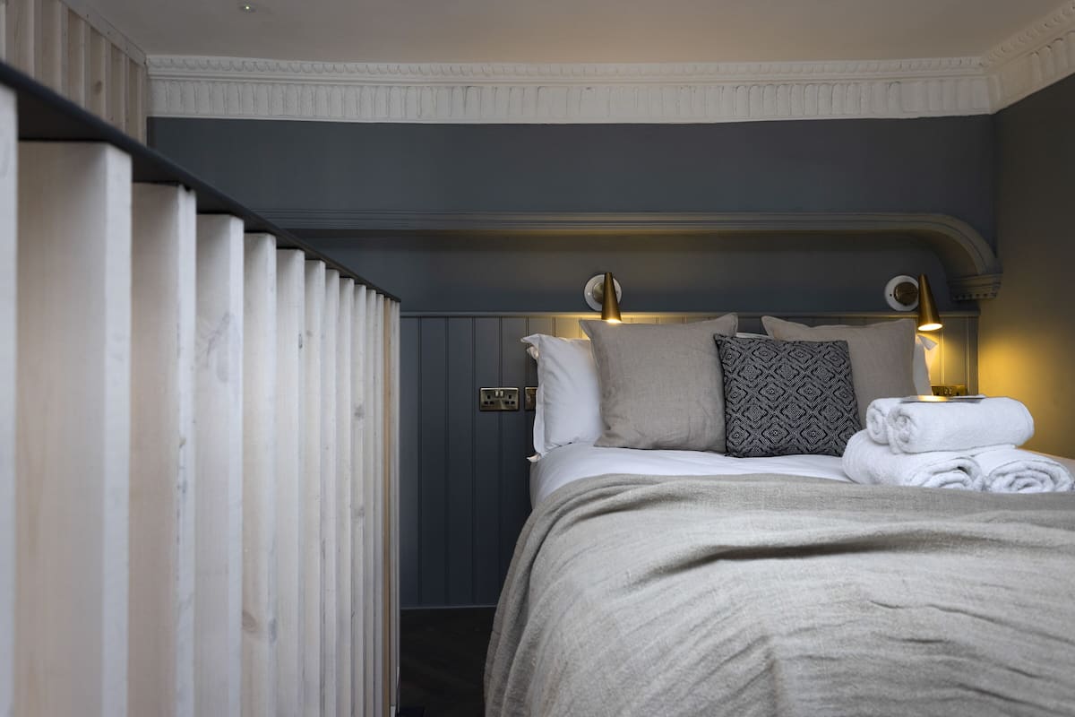 A neatly made bed with a beige blanket, a pillow, and a folded towel. There is a bedside lamp with a gold shade, and a wall socket is visible on the wall.
