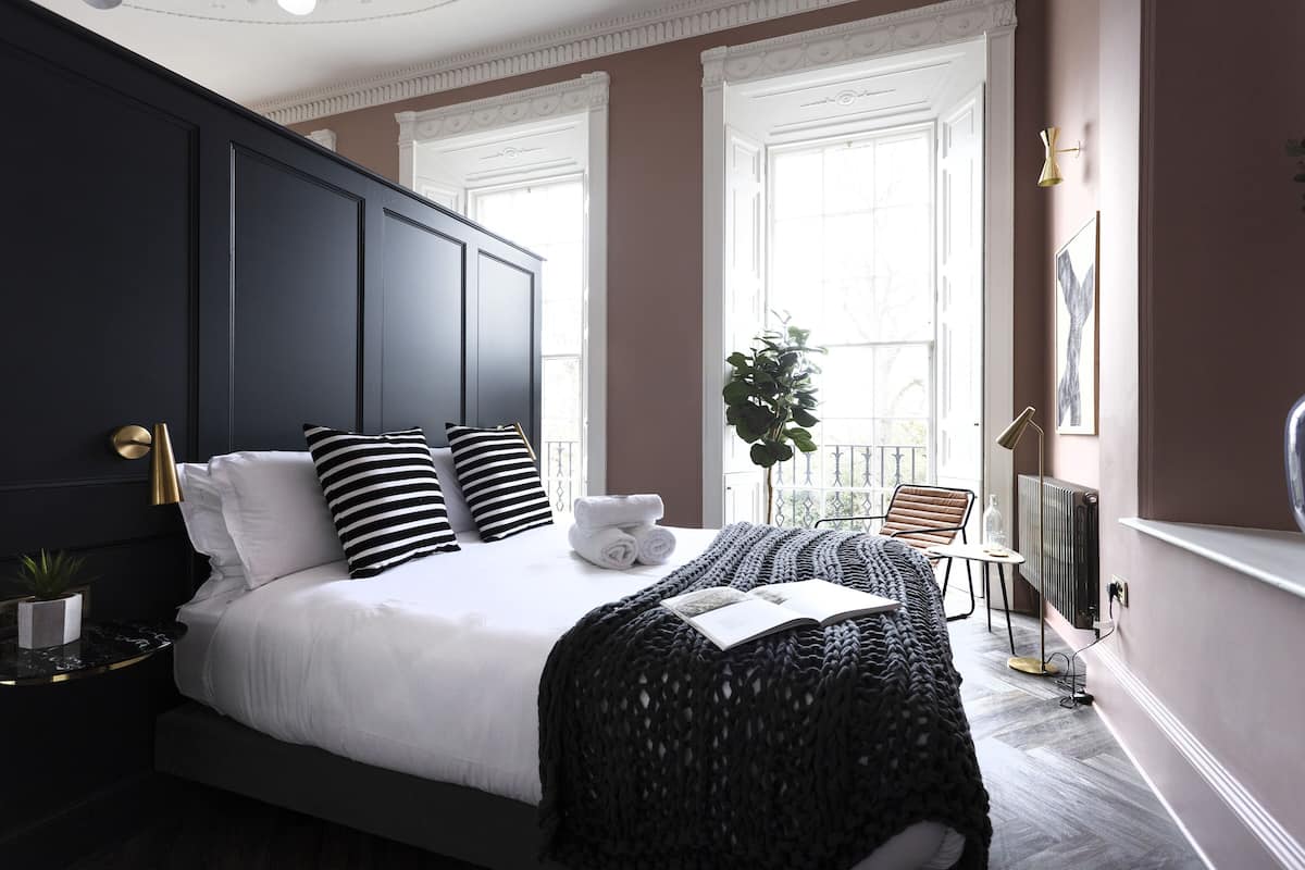 A modern bedroom with a large bed, a bedside table with a lamp, and a view of the outdoors through large windows.