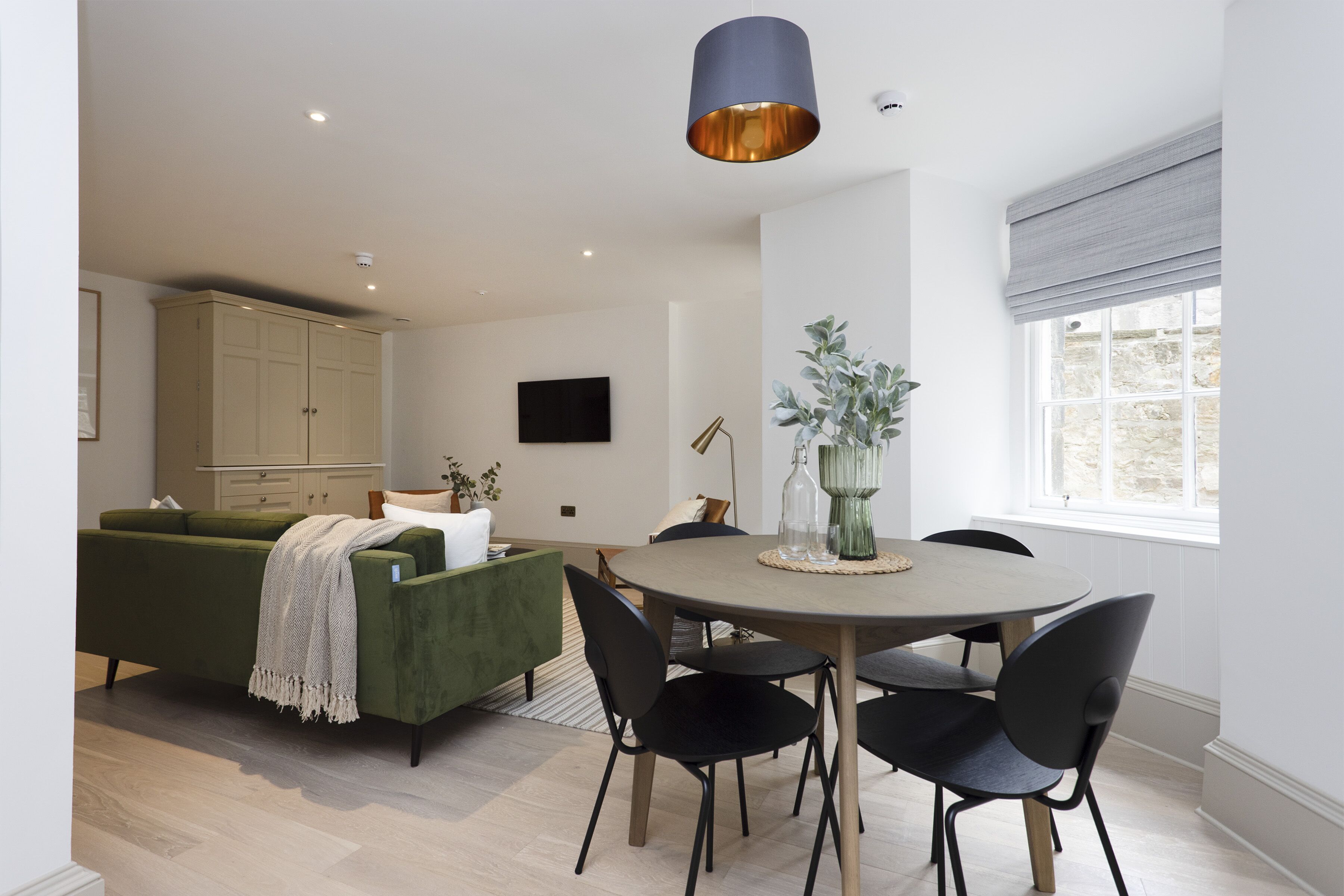 A modern living room with a green sofa, a round dining table with black chairs, a large window with a view, and a ceiling light fixture.