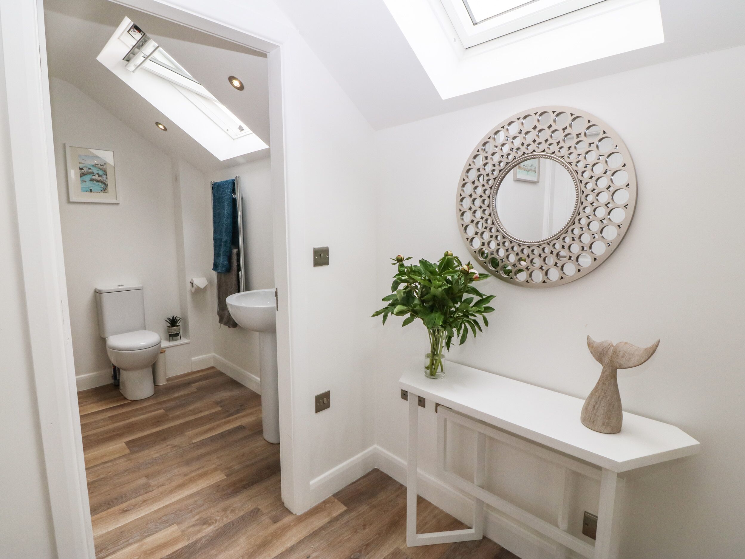 A modern bathroom with a skylight, a round mirror, a vase with green plants, and a wooden tail sculpture on a white console.
