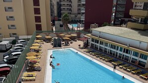 A rooftop pool area with sun loungers and umbrellas, surrounded by a high-rise building.