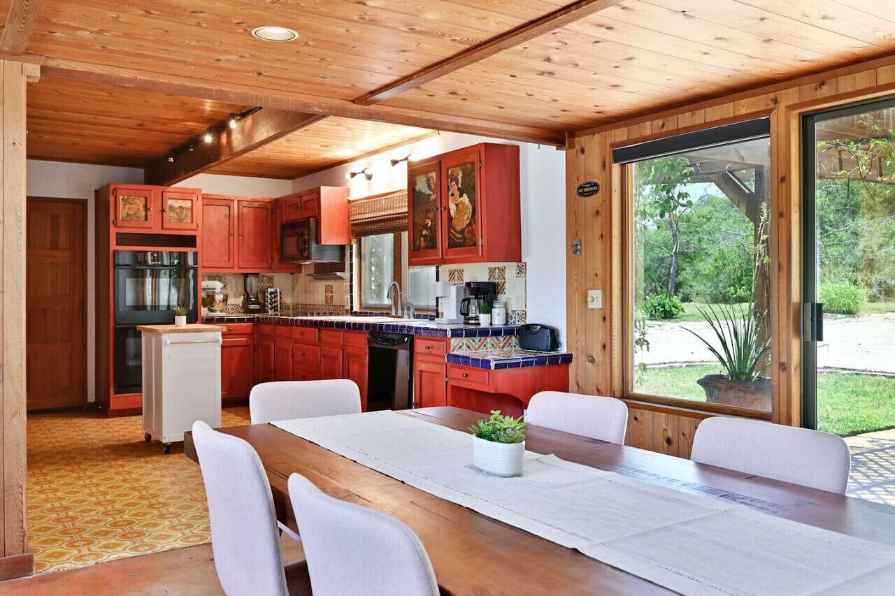 A kitchen with wooden cabinets, a dining table with chairs, and a view of the outdoors through large windows.