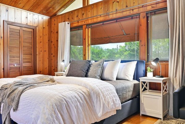 A bedroom with a large bed, a bedside table, a lamp, and a window with a view of trees.