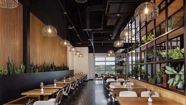 Ein modernes Restaurant mit Holztischen und -stühlen, hängenden Pendelleuchten und einer Wand mit Blumentöpfen.