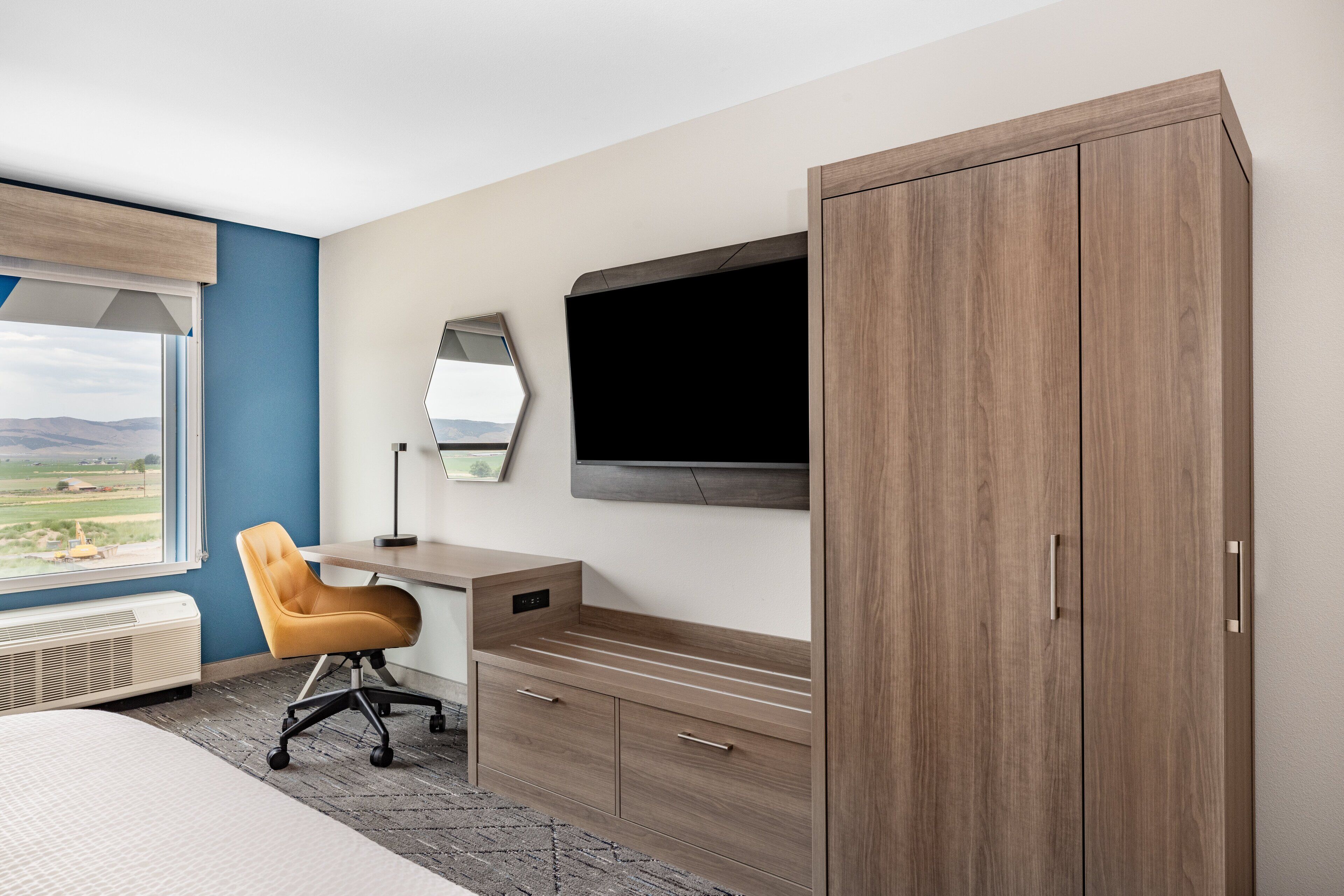 A bedroom with a bed, a desk, a chair, a wardrobe, a TV, and a window with a view of the countryside.
