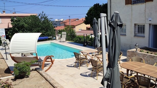 Zona de piscina con sillones y mesas, rodeada por un edificio con tejado de baldosas.